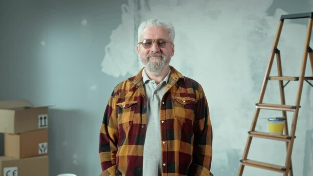 Elderly gray haired man with beard in glasses looking at camera and smiling. Portrait of pensioner in plaid shirt posing in an apartment against the background of a wall painted with white paint.