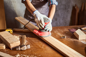 Female carpenter grinding wood with sandpaper in carpentry or diy workshop. Electric sander working in carpentry. Girl polishes wooden board with electric orbital sander.