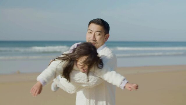 Happy father playing with his little daughter at seashore on autumn day. Asian man spinning cute girl around while child imagining flying like plane. Fun, parenthood, family concept