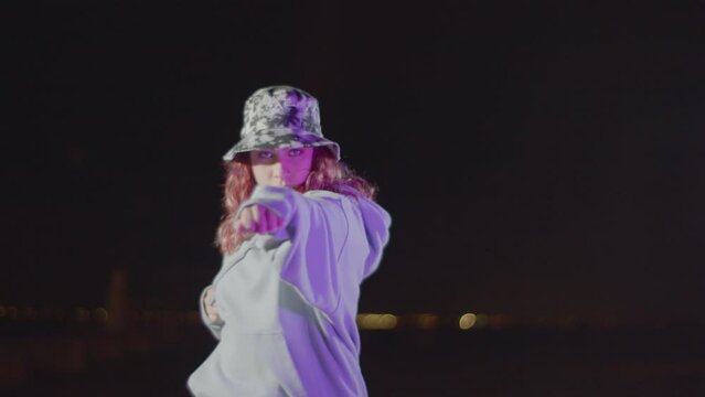 Pretty Caucasian woman standing outdoors against blurred city lights background, then turning to camera and making karate arm punch. Red-haired girl in camouflage hat showing dance move. Youth concept