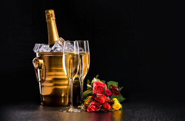 Two glasses of champagne, a bucket with a bottle of champagne and ice and a bouquet of multi-colored roses on a black background