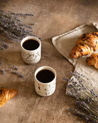 Two cups of black coffee with chocolate croissant on brown wood background with composition of lilac lavender flowers