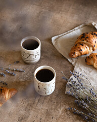 Two cups of black coffee with chocolate croissant on brown wood background with composition of lilac lavender flowers
