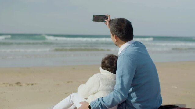 Cute little child spending time with her dad at seashore on sunny day. Man in blue jumper holding smartphone and taking selfie while hugging his lovely daughter. Leisure, family concept