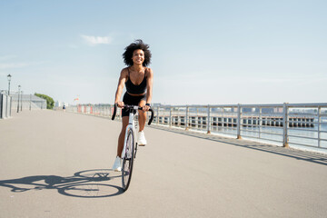 A young woman with curly hair in sports clothes riding a bicycle in the city. Ecological transport for every day. - obrazy, fototapety, plakaty
