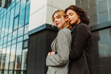 Two pretty women friends posing near glass building. Couple of gay lesbian girls hugging embracing together girlfriends, dressed casual outfits, have a date. LGBT concept. Fashion, make up, hairstyle

