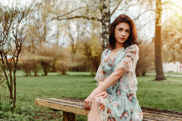 Fototapeta premium Portrait of charming pretty woman dressed flowery dress sitting on bench near apple cherry tree blossoms blooming flowers in the garden park in early spring nature. Fashion, holiday 