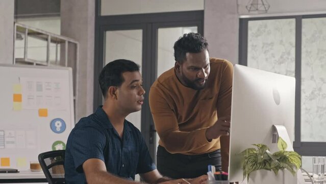 Indian IT specialist walking to colleague in office and giving him advice about project on computer while working together in open space office