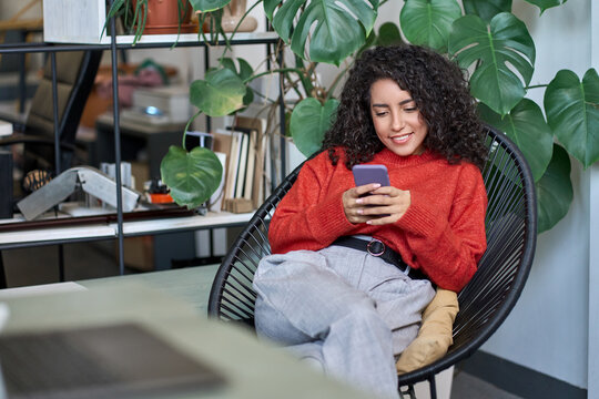 Young Smiling Business Woman Looking At Smartphone Using Cellphone Mobile Cellular, Professional Female Worker Or Student Typing On Mobile Cell Phone Sitting In Chair With Green Plants Around.