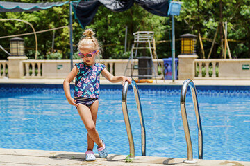 Happy little caucasian toddler girl with sunglasses smiling joyfully and enjoying summer vacations time in swimming pool in resort hotel. Kid with blonde hair wearing swimwear during summer holidays 