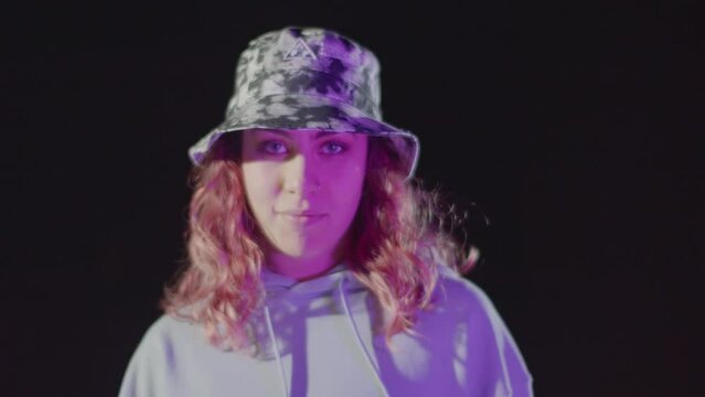 Portrait Shot Of Pretty Red-haired Young Lady Looking At Camera While Making Rhythmic Dance Moves Against Dark Background. Female Dancer In Camouflage Hat Smiling Mysteriously. Attitude, Youth Concept