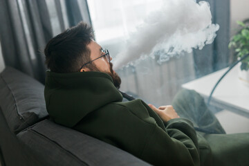 Bearded millennial or gen z man smoking hookah while relaxing on sofa at home - chill time and...