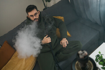 Bearded millennial or gen z man smoking hookah while relaxing on sofa at home - chill time and resting concept