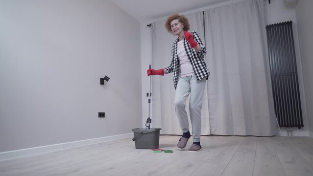 Senior woman is cleaning up before moving into a new apartment dancing and singing along to my floor with a mop. Excited woman enjoying housekeeping. People, housework and housekeeping concept. 