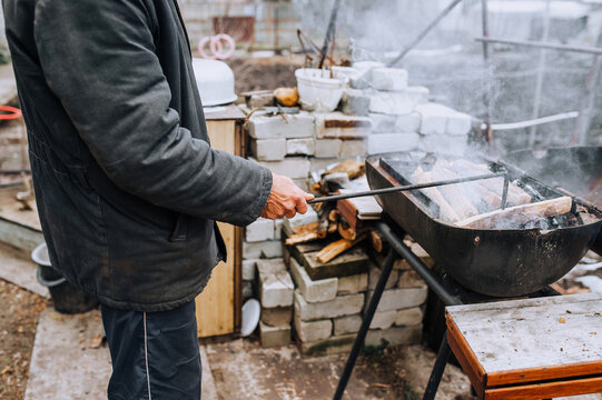 A man outdoors, in nature, holds a metal poker in his hands and corrects burning firewood in a barbecue with smoke. Photography, food preparation.