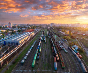 Aerial view of freight trains at sunset. Colorful railway cargo wagons on railroad. Drone view of wagons, city, sky with orange clouds. Depot of freight trains. Railway station. Industrial landscape