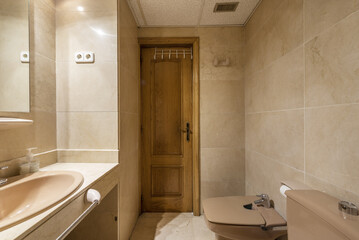 Bathroom with marble tiles, thick marble countertop with embedded porcelain sink, frameless mirror, brown toilets and oak wood door