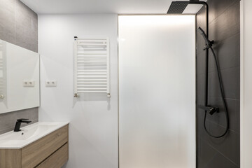 Newly renovated bathroom with black taps, square mirror, black tiles in the shower, white heated towel rail and light wood furniture with drawers