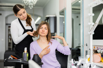 Interested female client sitting in chair while discussing haircut with positive girl professional hairdresser ..