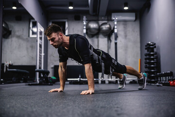 Obraz na płótnie Canvas Modern training concept on electrical muscle stimulation. Side view of a male wearing an EMS suit and doing arm exercises and push-ups. He is in a plank position and he touching the floor with hands