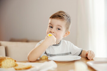 A hungry little boy is sitting at the dining table at home and eating boiled egg for a breakfast.