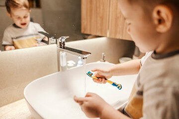 A little boy is washing his toothbrush in sink in the bathroom in the morning.