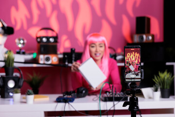 Fototapeta na wymiar Asian woman with pink hair holding white box advertising product while recording vlog using smartphone camera in broadcast studio. Influencer filming content using professional equipment