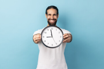 Portrait of happy man with beard wearing white T-shirt holding out wall clock, being happy,...