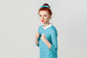 Portrait of confident serious red haired woman with hair bun, standing with clenched fists, being ready to attack, boxing, wearing blue dress. Indoor studio shot isolated on gray background.