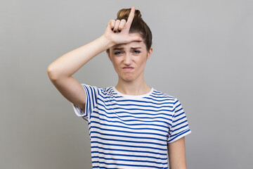 Portrait of upset disappointed woman wearing striped T-shirt standing, showing looser gesture with hand on forehead, looking at camera. Indoor studio shot isolated on gray background.