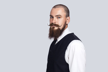 Side view portrait of serious attentive man with beard and mustache standing looking at camera with strict facial expression, being in bad mood. Indoor studio shot isolated on gray background.