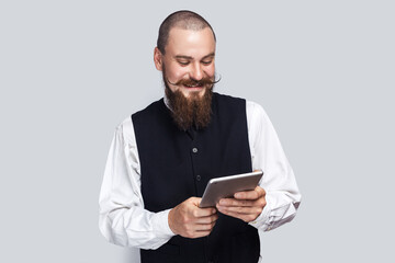Portrait of extremely happy positive man with beard and mustache using tablet and laughing, reading jokes or watching funny video. Indoor studio shot isolated on gray background.