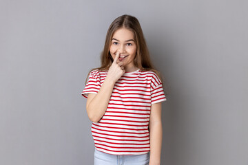 Portrait of weird little girl wearing striped T-shirt holding finger in her nose showing tongue, uncultured bored girl having fun, bad manners. Indoor studio shot isolated on gray background.