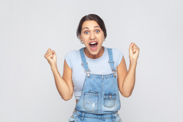 I am champion. Portrait of victorious delighted woman wearing denim overalls clenching fists, shouting for joy, screaming celebrating win success. Indoor studio shot isolated on gray background.