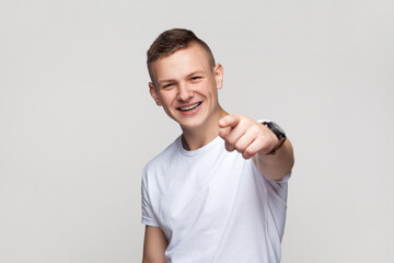 Portrait of satisfied smiling positive young teenager boy in T-shirt looking at camera with toothy smile, pointing at camera with finger, choosing you. Indoor studio shot isolated on gray background.