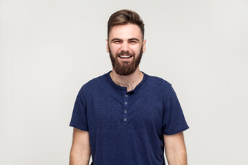 Obraz premium Portrait of overjoyed extremely happy handsome man with beard wearing dark blue T-shirt hunching from laughter, laughing out loud, crazy face. Indoor shot isolated on gray background.