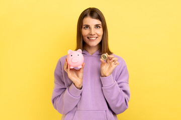 Fototapeta na wymiar Portrait of young adult happy smiling pretty woman with hair holding piggy bank and gold crypto coin, growth bitcoin, wearing purple hoodie. Indoor studio shot isolated on yellow background.