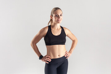 Fototapeta na wymiar Self confident sporty blonde woman standing and looking at camera, keeps hands on hips, posing after workout, wearing black fitness clothing. Indoor studio shot isolated on gray background.
