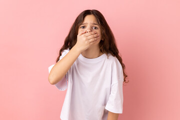I won't tell. Portrait of cute little girl wearing white T-shirt covering mouth with hand, keeping secret, terrified with shocking news. Indoor studio shot isolated on pink background.