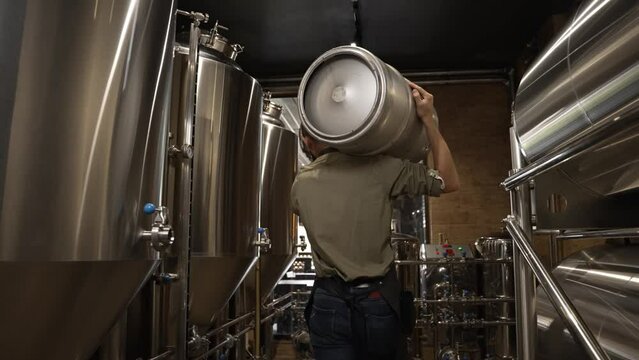 A working male brewer carries a keg filled with beer as he passes beer tanks