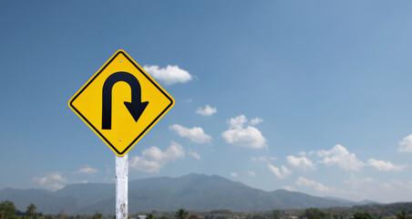 Traffic sign: Right U-turn sign on cement pole beside the rural road with white cloudy bluesky and mountains background, copy space.	