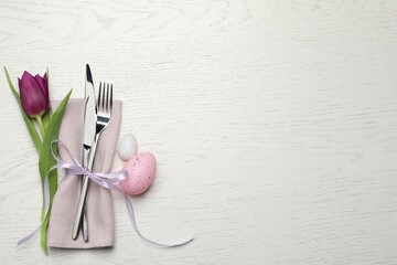 Cutlery set, painted eggs and beautiful flower on white wooden table, flat lay with space for text. Easter celebration