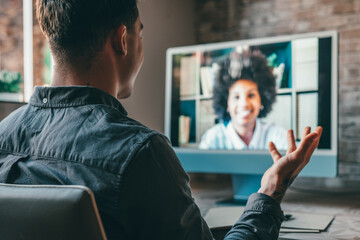 View over shoulder employer listen applicant at job interview online use cam and pc. Young caucasian man with tattoos at home working and talking online with African woman colleague in videocall.