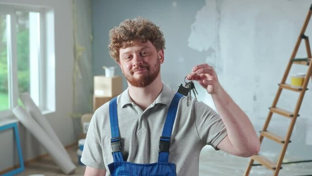 Repairman worker in blue construction overalls is showing bunch of keys and smiling. Portrait of redhead man is posing against backdrop of apartment in process of renovation, ladder, cardboard boxes