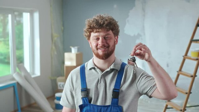 Repairman worker in blue construction overalls showing bunch of keys and smiling. Portrait of redhead man posing against backdrop of apartment in process of renovation, ladder, cardboard boxes, window