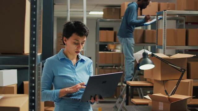 Female worker checking cardboard packages from storage room racks, using laptop to plan stock inventory and take notes. Entrepreneur working on business development and supply chain.