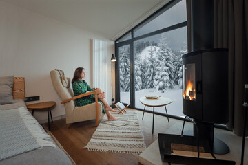 Woman resting in cozy house with fireplace near window with winter panoramic view