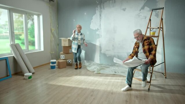 Elderly man and woman are looking through sheet with plan of an apartment and discussing renovation project. Aged couple is having fun and celebrating good start to renovation.