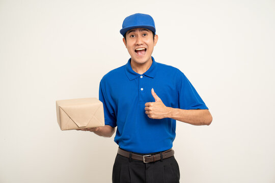 Happy delivery asian man in blue uniform standing holding box parcel cardboard on isolated white background. Smiling male service worker. Delivery courier and shipping service.