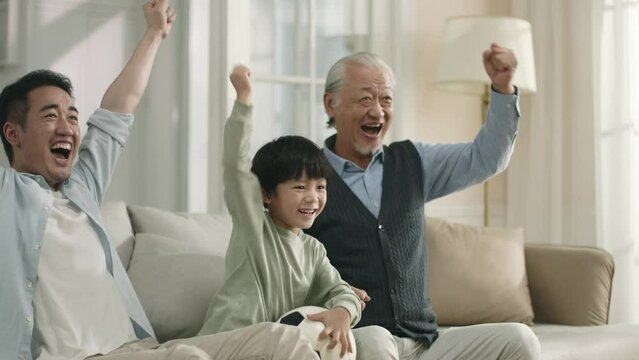 asian grandfather father and son sitting on family couch watching football game on tv at home